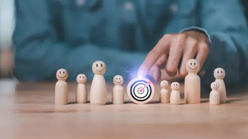 person sitting behind small wooden figures with smiling faces, holding his finger on a wooden target in the center.